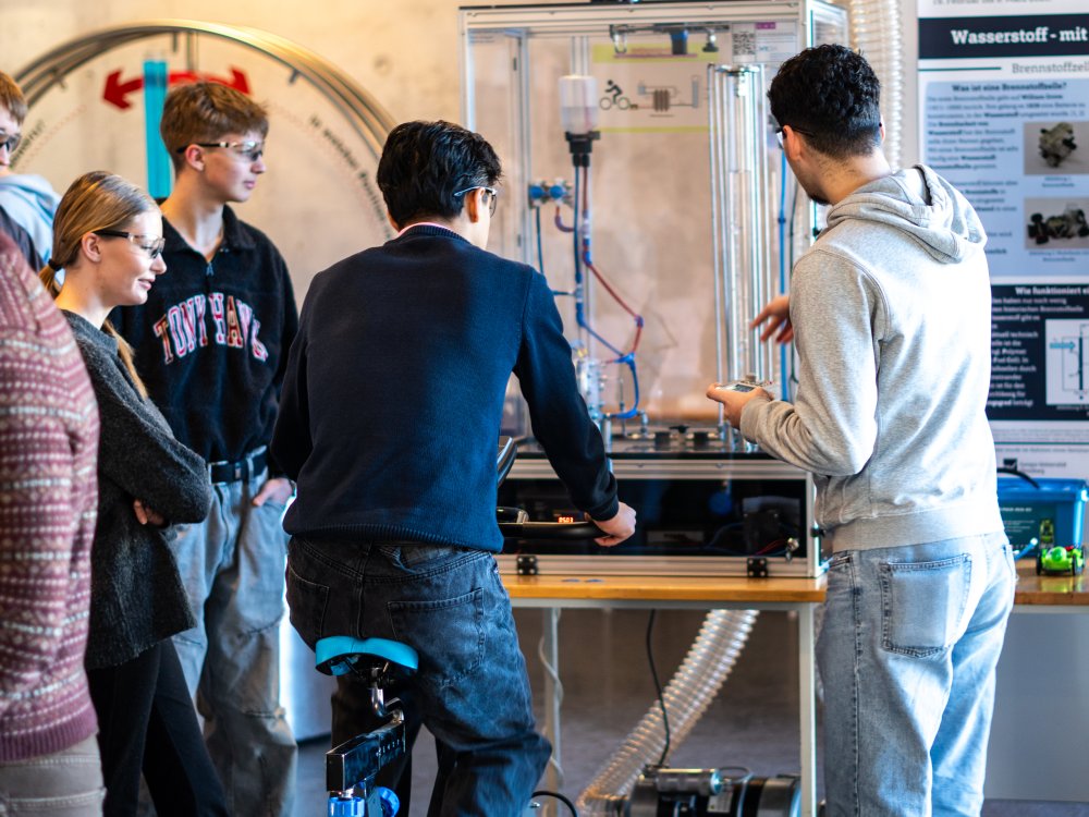 Ein junger Mann sitzt auf einem Fahrrad-Ergometer und tritt in die Pedalen, drei weitere Schüler schauen zu, wie er dabei durch Elektrolyse Wasserstoff produziert.
