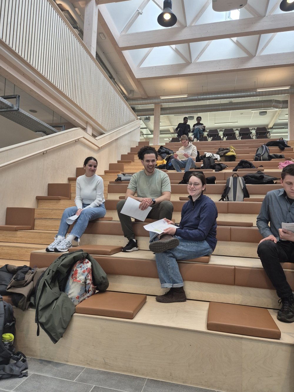 Auf einer Treppe in einem hellen Atrium sitzt eine Gruppe von Studierenden und lacht in die Kamera