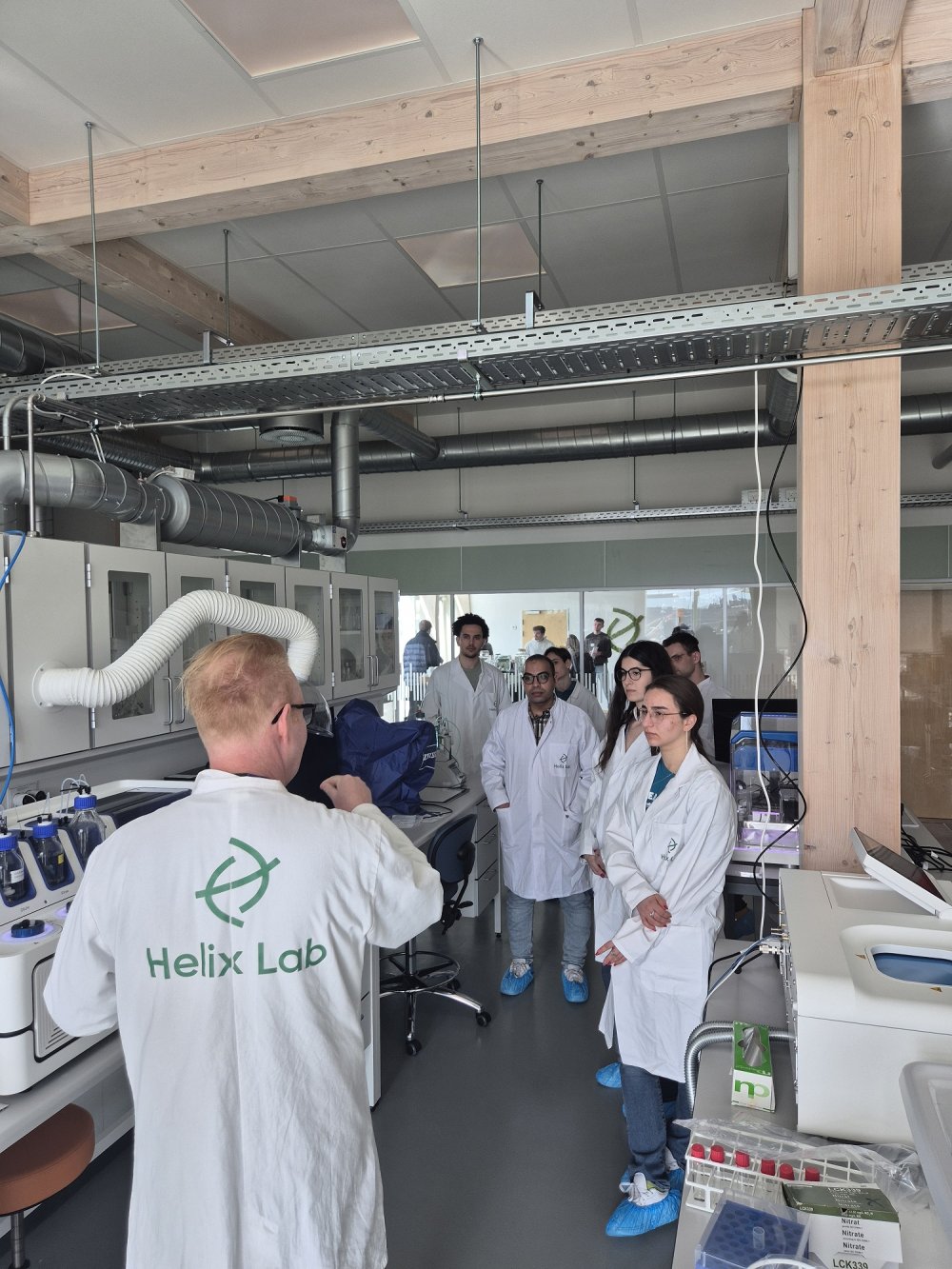 In einem Labor steht eine Gruppe von Studierenden in Laborkitteln, einer von ihnen hat den Rücken zur Kamera und hat auf dem Rücken die Aufschrift &quot;Helix Lab&quot;