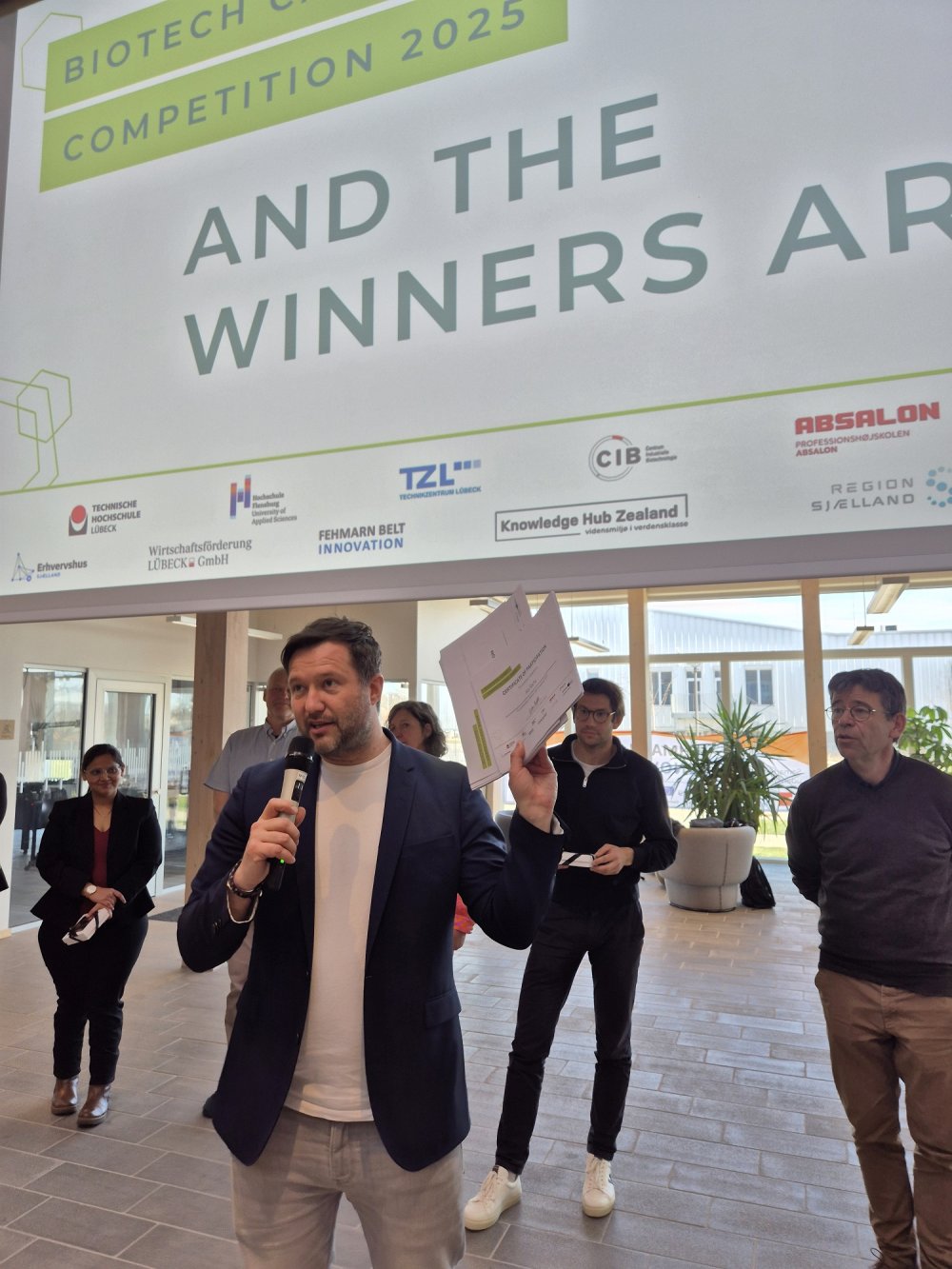 Ein Mann spricht auf einer Bühne in ein Mikro, hinter ihm eine Leinwand mit Text: And the winners are