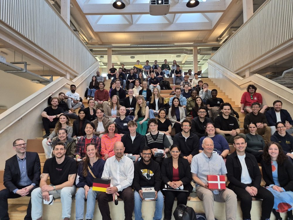 Eine große Gruppe von mehr als 50 Studierenden sitzt auf einer Treppe in einem hellen Raum