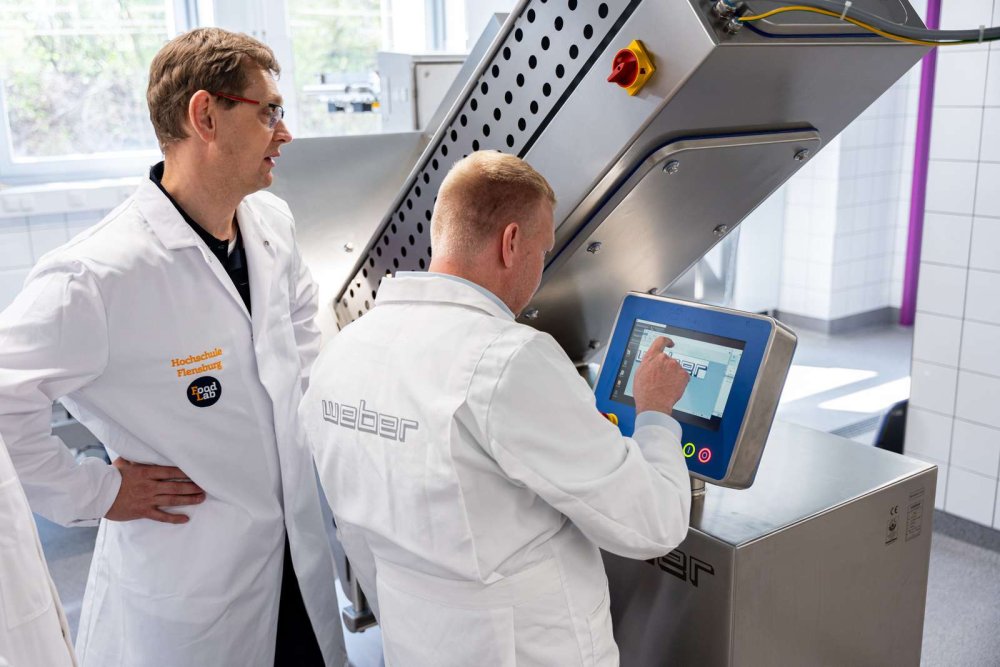 Zwei Männer mittleren Alters in Laborkitteln stehen an einem großen silbernen Laborgerät und machen Eingaben an einem Monitor.