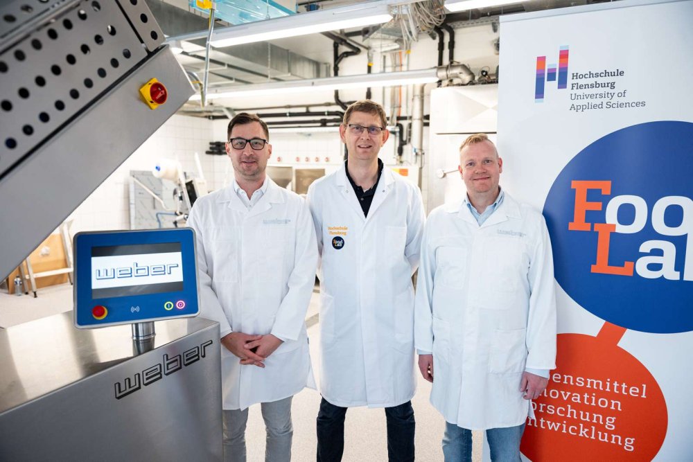 Drei Männer mittleren Alters in Laborkitteln stehen in einem Labor zwischen einem großen silbernen Gerät und einem Roll-Up mit dem Aufdruck Food Lab.