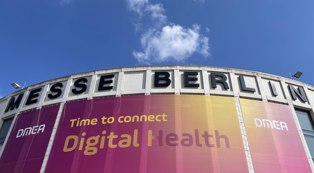 Blick aus Froschperspektive auf ein Gebäude der Messe Berlin mit einem großen lila-orangenen Banner: DMEA - Time to connect - Digital Health