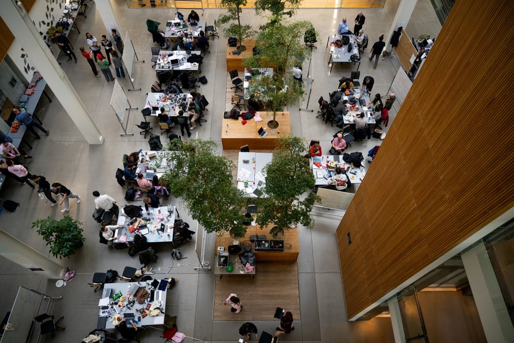 Blick von oben in einen weiten, offenen Raum mit vielen Tischen, an denen Gruppen von Studierenden zusammensitzen. Im Raum verteilt sind mehrere kleine Bäume.