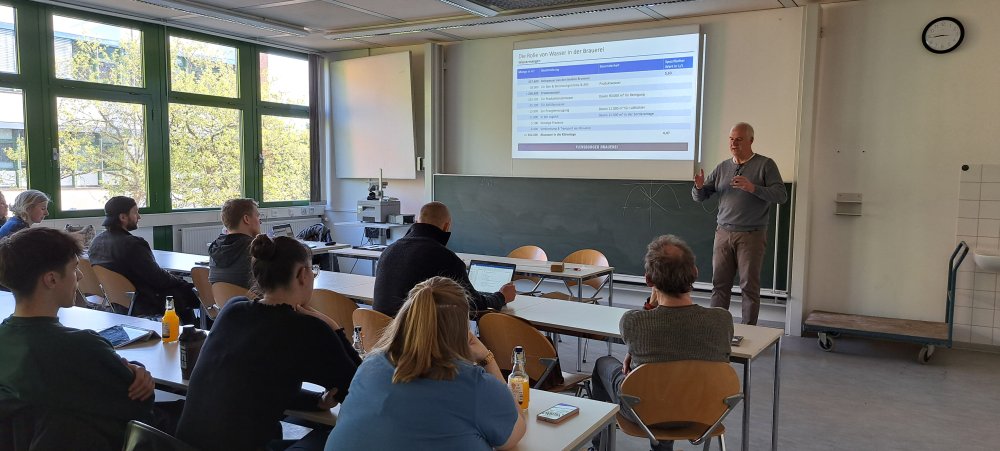 In einem Seminarraum sitzen Studierende, vorne steht ein Mann und hält einen Vortrag, seine Präsentationsfolie trägt den Titel &quot;Die Rolle von Wasser in der Brauerei&quot;.