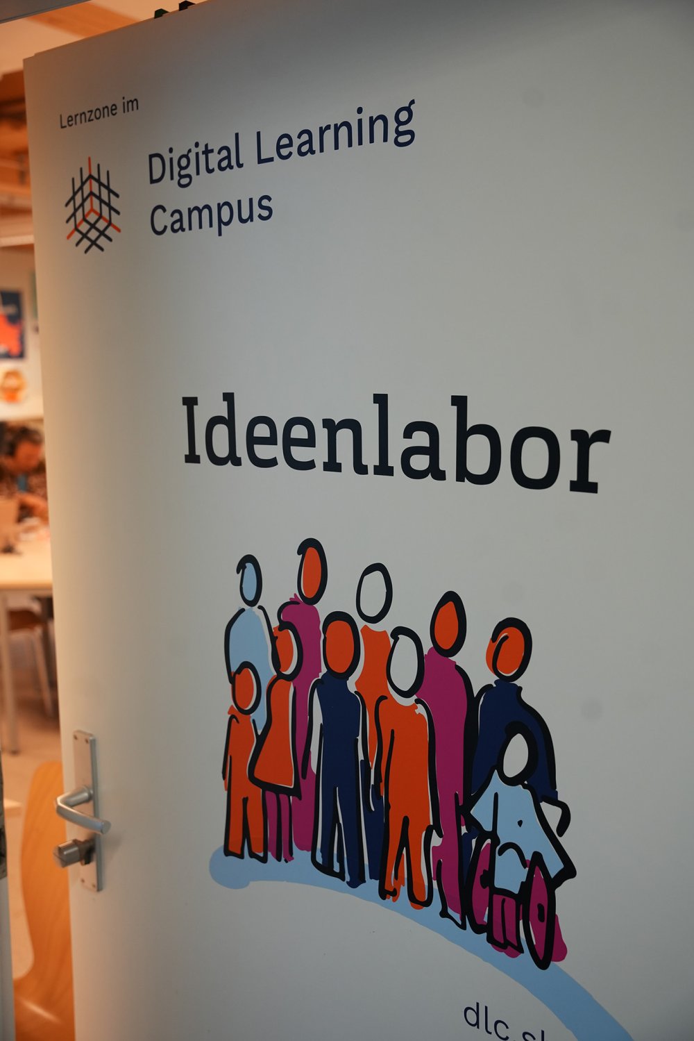 Offen stehende Tür zu einem Raum mit Schriftzug "Digital Learning Campus - Ideenlabor" und einer bunten Zeichnung von einer Menschengruppe.