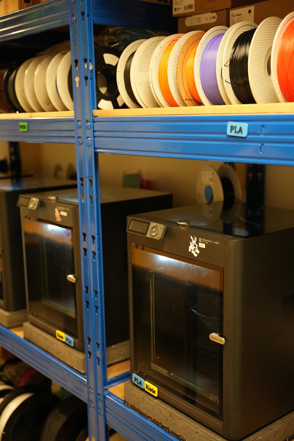 Schwerlastregal mit 3D-Druckern und Rollen mit bunten Bändern.