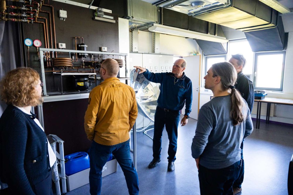 Eine Gruppe von fünf Menschen steht in einem Raum vor einer Brennstoffzellen-Anlage, ein Mann erklärt die Anlage. Im Hintergrund steht ein großer Parabolspiegel.