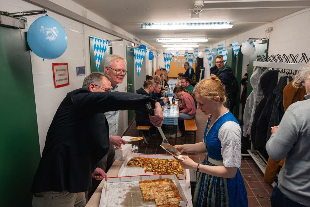 In einem Raum sitzen zahlreiche Menschen an Bierbänken, im Vordergrund wird Pizza ausgegeben.