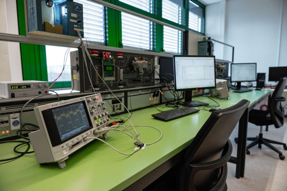 Auf dem Bild ist ein Beispiel für einen Arbeitsplatz im Laborraum zu sehen. Auf dem Tisch stehen ein Monitor, ein Oszilloskop und weitere Laborausstattung.