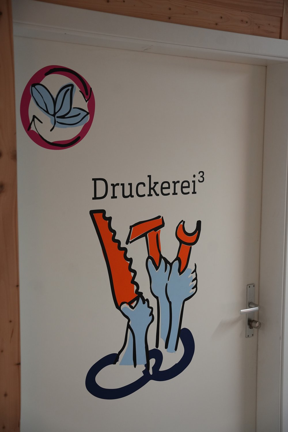 Tür mit buntem Aufdruck "Druckerei³" und mehreren Händen, die Werkzeuge halten.