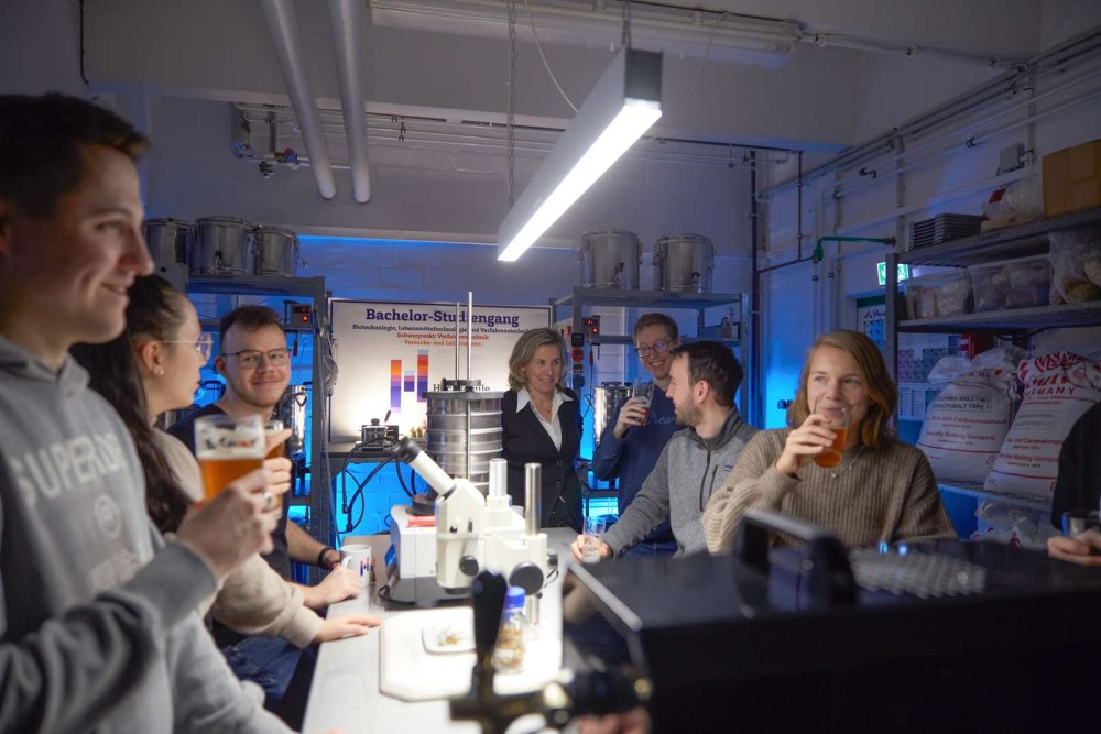 In einem Laborraum stehen Studierende um einen Tisch, unterhalten sich und trinken Bier.