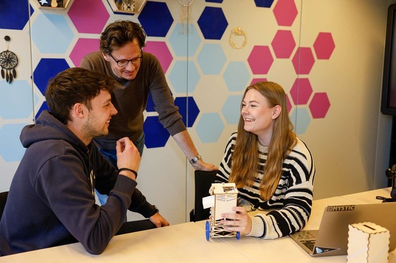 Zwei Studierende, ein junger Mann und eine junge Frau, sitzen in einem freundlichen Arbeitsraum an einem Tisch, neben ihnen steht ein Dozent. Die Studentin hält einen kleinen Roboter in der Hand.