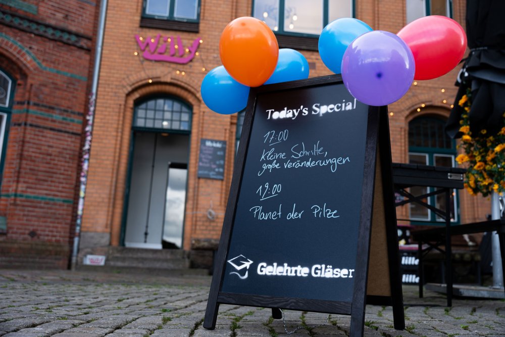 Aufstell-Tafel vor einem Lokal. Darauf Hinweise auf zwei Vorträge, an der Tafel sind bunte Luftballons festgebunden.