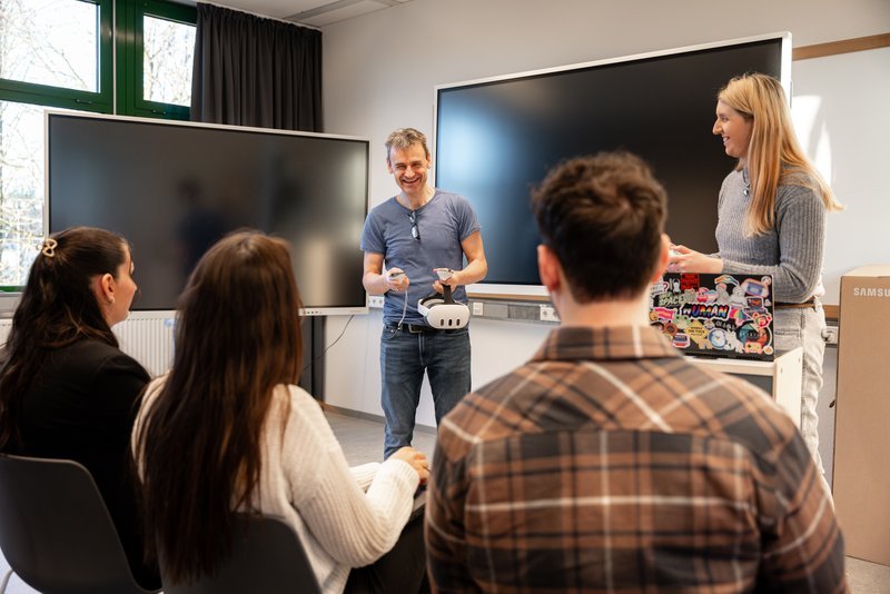 Ein lachender Mann mit VR Brille in der Hand steht vor Studierenden