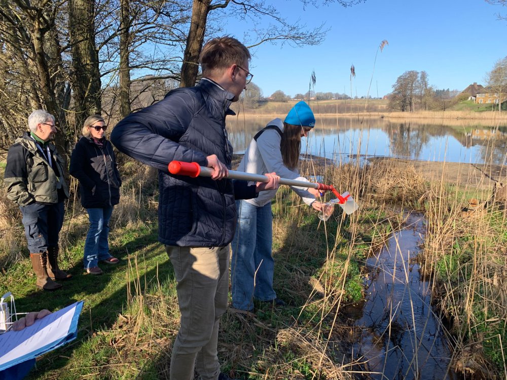 Studierende neben eine Wasserprobe. Sie stehen an einem bachartigen Zufluss zu einem See, der im Hintergruind zu erkennen ist.