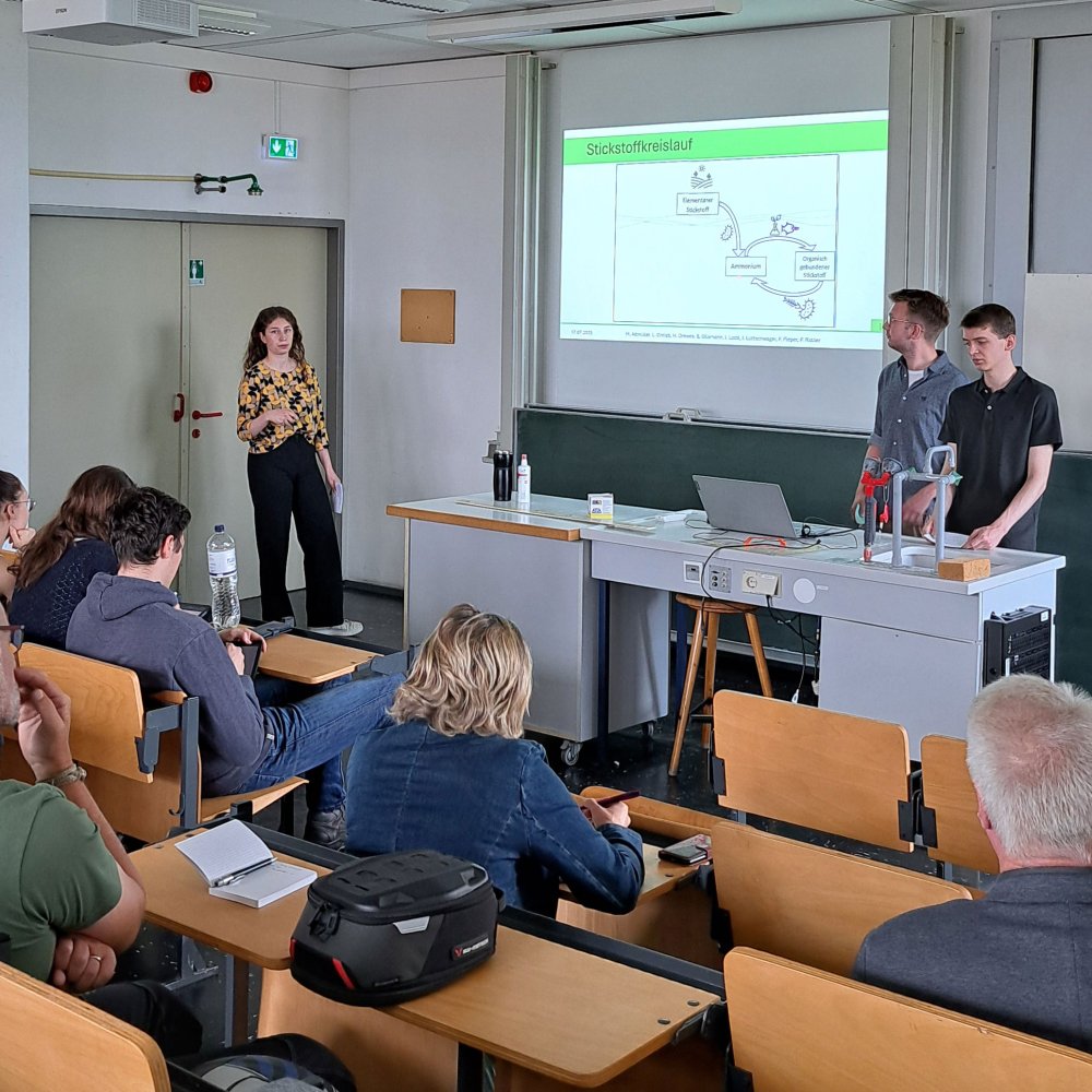 Präsentationssituation: 3 Studierende vor einer Gruppe Menschen in einem Hötrsaal. Sie deuten auf eine an die Wand projizierte Präsentation.
