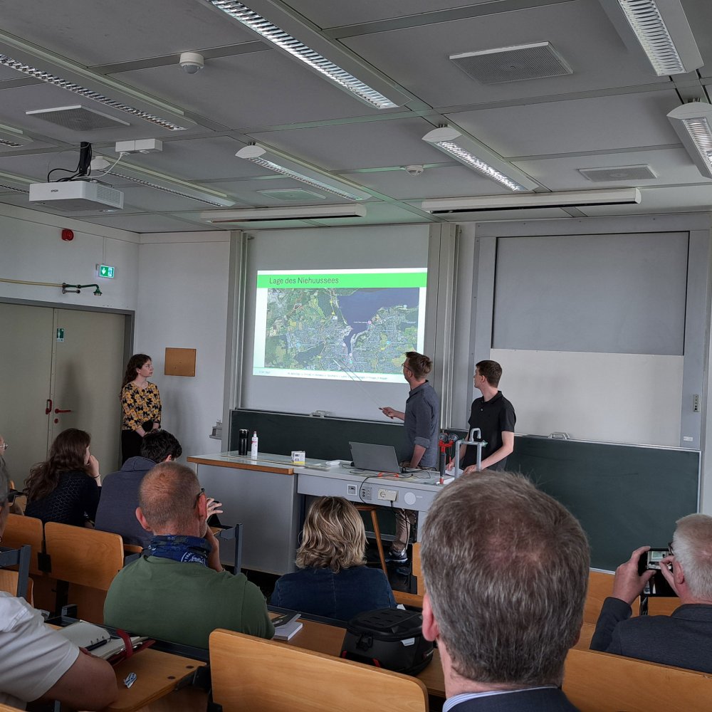 Präsentationssituation: 3 Studierende vor einer Gruppe Menschen in einem Hötrsaal. Sie deuten auf eine an die Wand projizierte Präsentation.