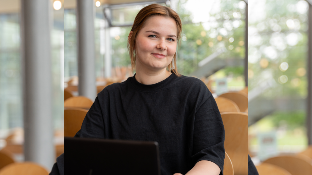 Junge Frau in schwarzem Oberteil sitzt in der Flensburger Mensa an einem Laptop und schaut in die Kamera.