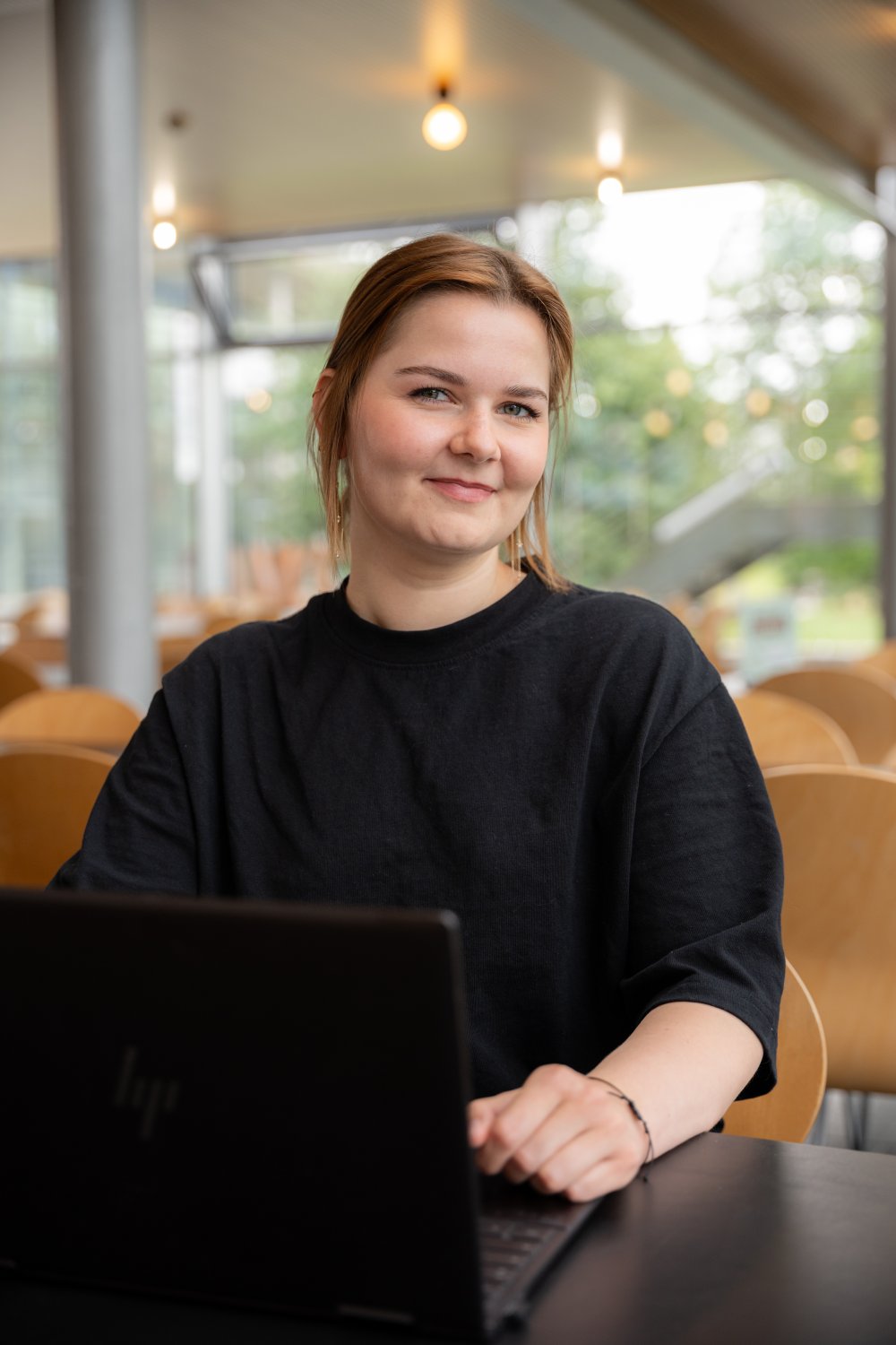 Junge Frau in schwarzem Oberteil sitzt in der Flensburger Mensa an einem Laptop und schaut in die Kamera.