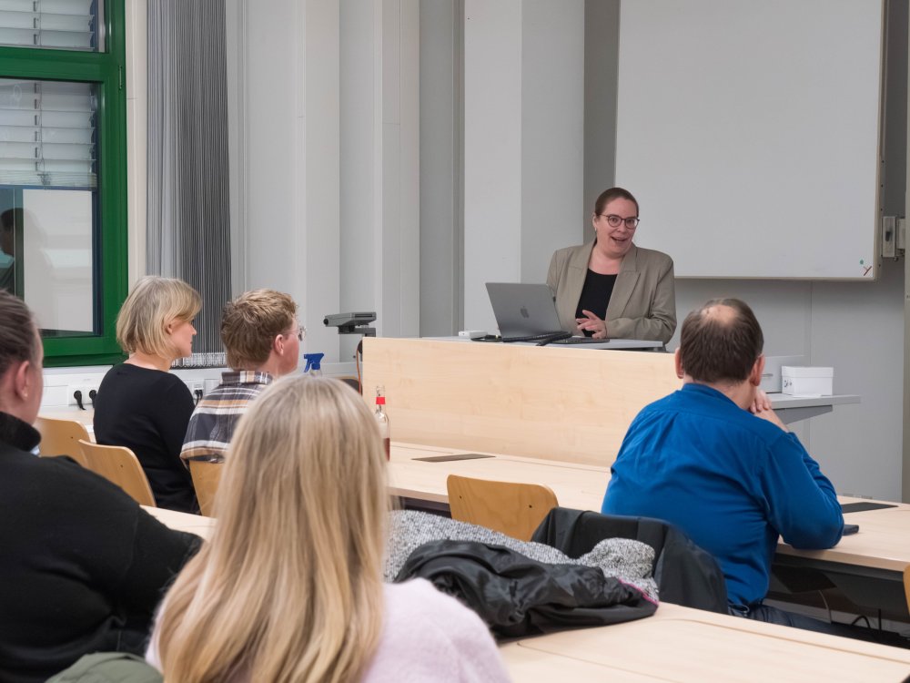 Eine Frau steht am Rednerpult, viele Menschen, mit dem Rücken zum Betrachter, hören zu