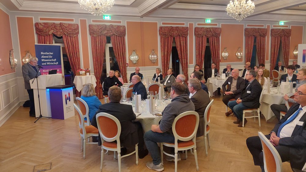 Ein Saal mit runden Tischen an denen Menschen sitzen, die einem Mann zuhören, der an einem Rednerpult steht und spricht