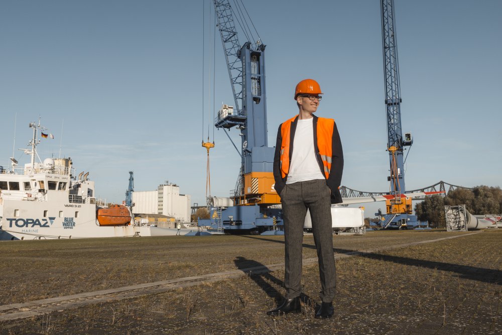 Ein Mann mit oragener Weste und orgenemen Helm steht vor Hafenanlagen