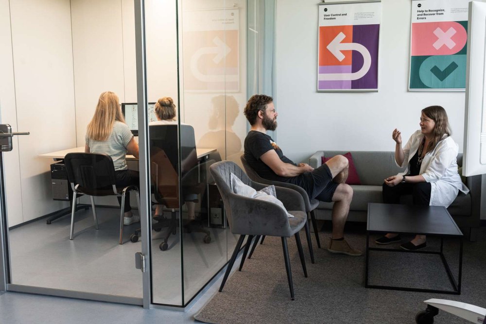 In einem Laborraum sitzen zwei junge Frauen an einem PC in einem mit Glas abgetrennten Arbeitsbereich, daneben sitzen eine Frau und ein Mann in einer Sitzecke mit Sofa und unterhalten sich.