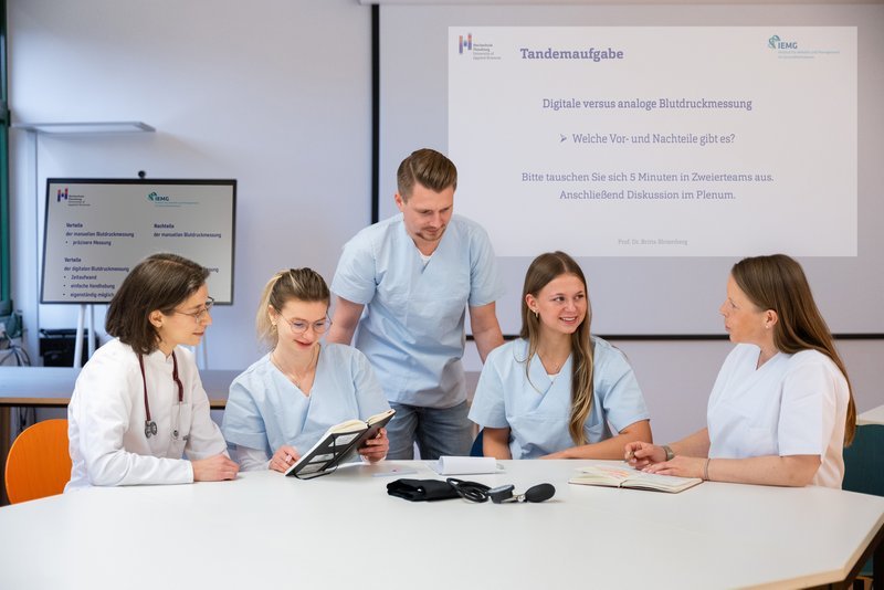 fünf Menschen sitzen am Tisch, einer steht, sie unterhalten sich, haben weiße Kittel an