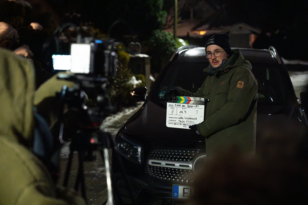 Ein Studierender mit einer Filmklappe vor einem Auto.