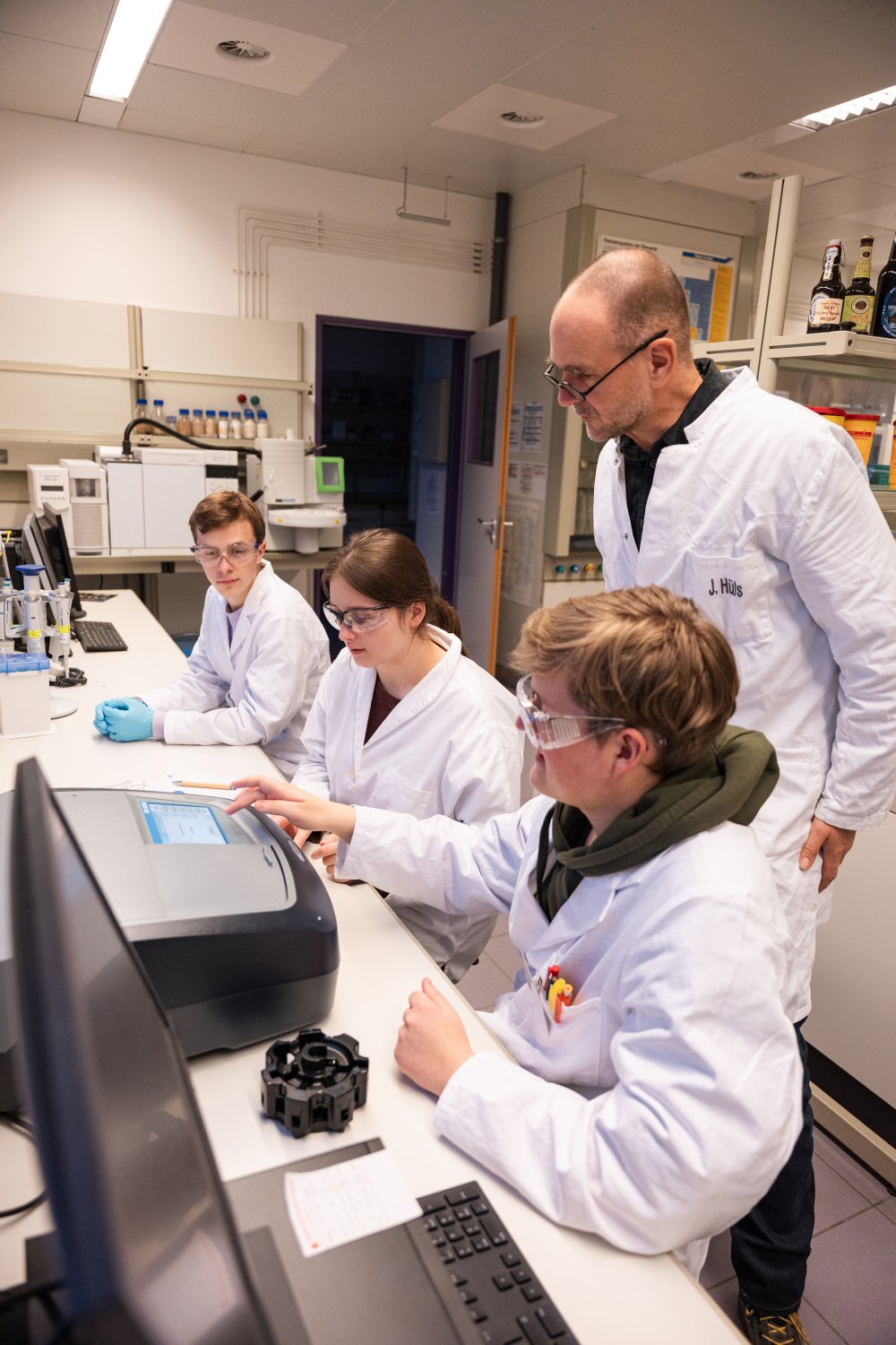 Ein mann steht hinter jungen Menschen im Labor in weißen Kitteln und beobachtet, was sie machen