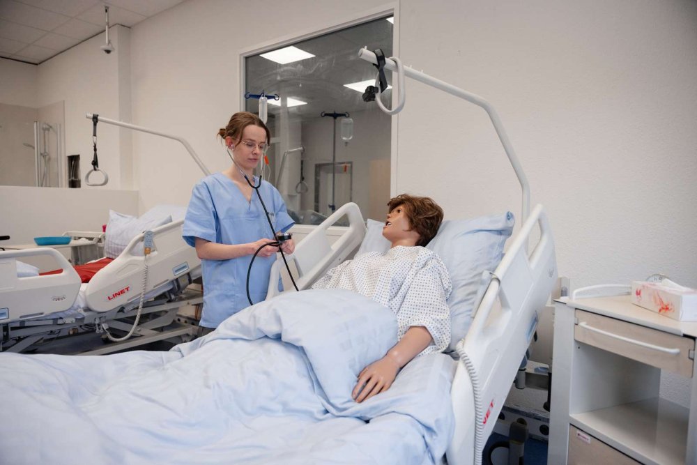Eine Studentin steht an einem Krankenhausbett, in dem eine lebensgroße Puppe liegt, und übt die Blutdruckmessung.