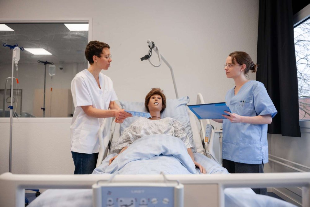 Eine Dozentin und eine Studentin stehen an einem Krankenhausbett, in dem eine lebensgroße Puppe liegt.