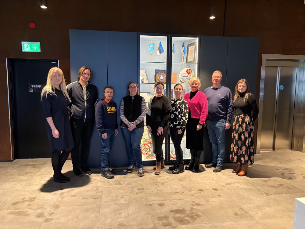 Eine Gruppe von Menschen steht in einem Foyer vor einer Vitrine mit estnischen Flagen und Kunst.