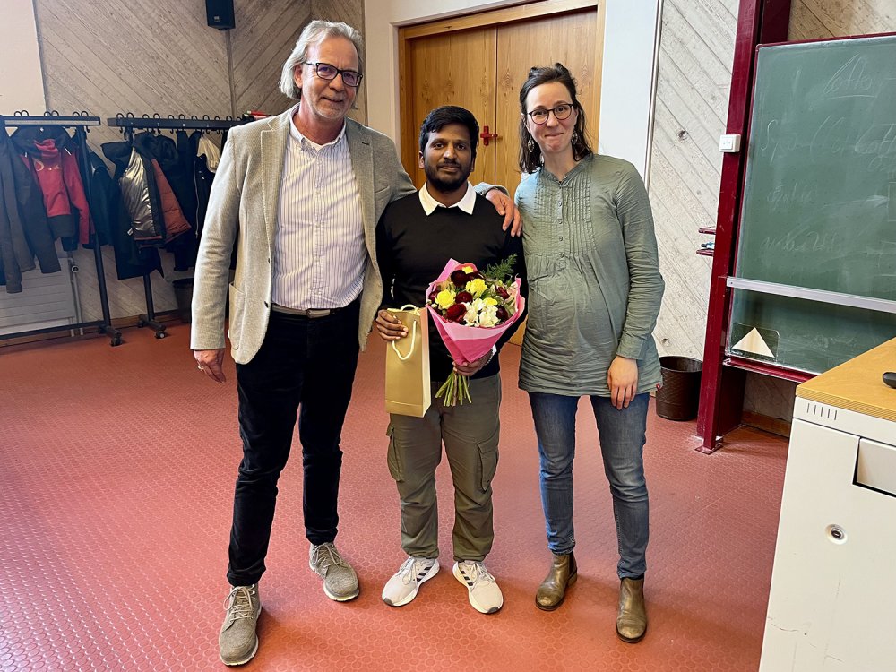 Ein älterer Mann im Sakko, ein junger Mann und eine junge Frau stehen zusammen in einem Vorlesungssaal und lächeln in die Kamera.