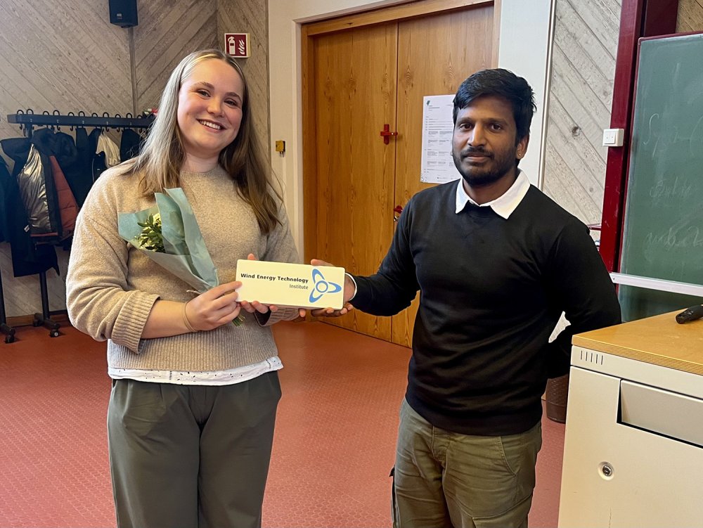 Eine junge Frau mit einem Blumenstrauß und ein junger Mann halten ein Schild mit dem Aufdruck "Wind Energy Technology Institute".