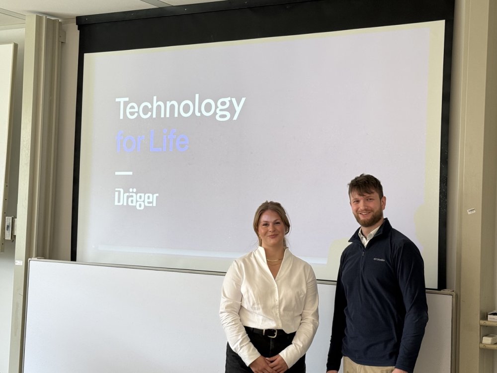 Eine junge Frau in weißer Bluse und ein junger Mann im blauen Zip-Pulli stehen vor einer Präsentationsfolie mit Text: Technology for Life - Dräger.