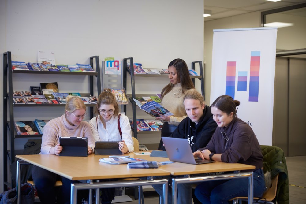 Fünf Studierende arbeiten gemeinsam an einem Tisch mit Laptop, Tablet und Broschüren. Die Szene zeigt, wie Lernen an der Hochschule zunehmend digital, kollaborativ und medienübergreifend stattfindet.