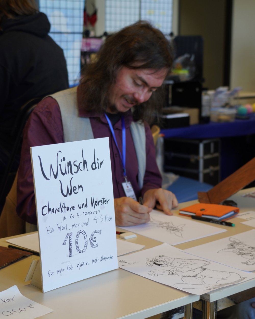 Messestand, an dem ein Mann mit langen Haaren sitzt und Comicfiguren zeichnet.