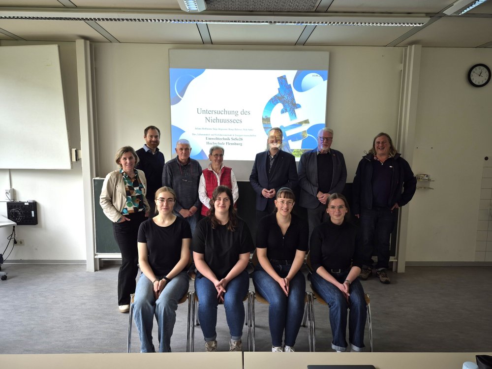 Eine Gruppe von Studierenden sitzt in einem Seminarraum, hinter ihnen stehen einige Männer und Frauen mittleren Alters vor einer Präsentation mit dem Titel "Untersuchung des Niehuussees".