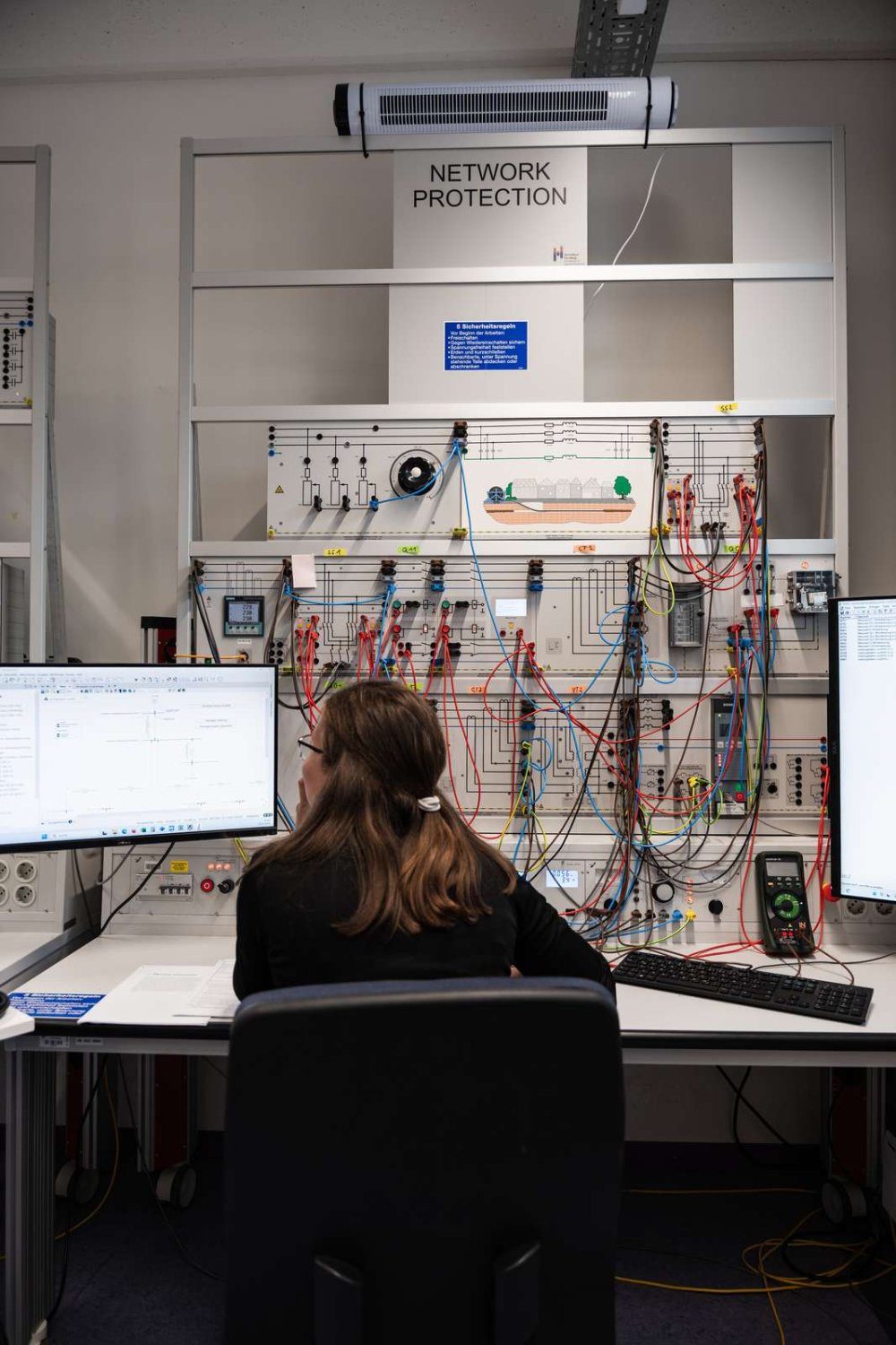 Eine junge Frau sitzt an einem PC-Arbeitsplatz mit einer elektrischen Schaltwand.
