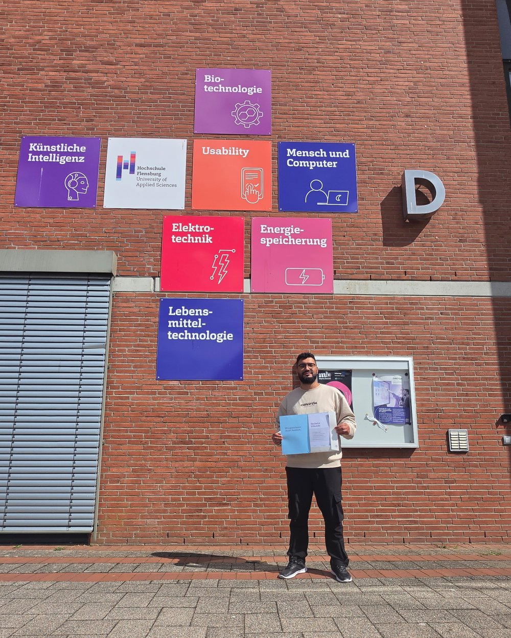 Ein junger Mann steht mit einer Urkundenmappe vor dem D-Gebäude der Hochschule Flensburg.