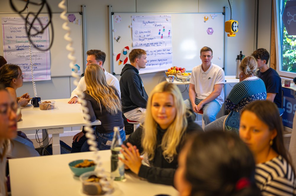 Junge Leute sitzen an mehreren Tischen in einem Seminarraum verteilt und unterhalten sich.