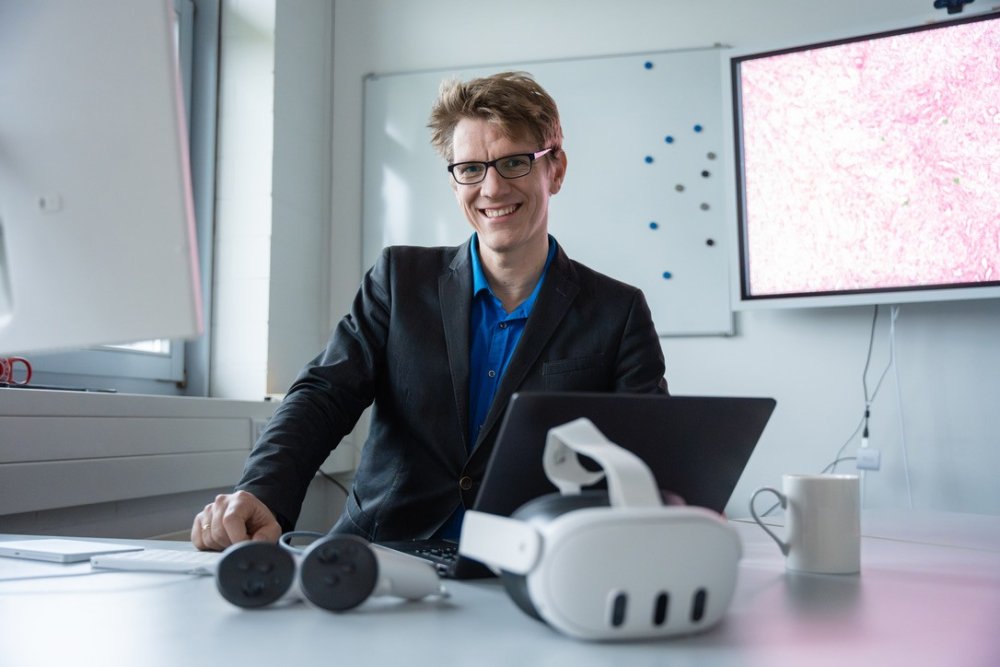 Person an einem Arbeitsplatz mit Laptop und VR-Brillen; im Hintergrund zeigt ein Bildschirm eine stark vergrößerte medizinische Gewebeaufnahme.