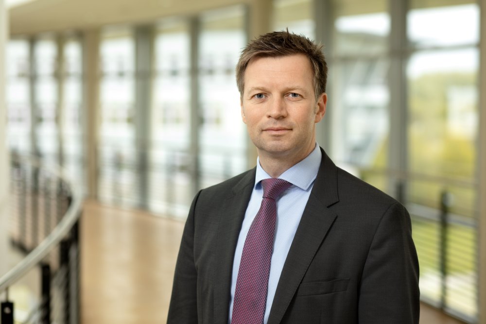 Porträt von Prof. Dr. Ilja Tuschy im Anzug vor einer hellen Fensterfront im Gebäude der Hochschule Flensburg.
