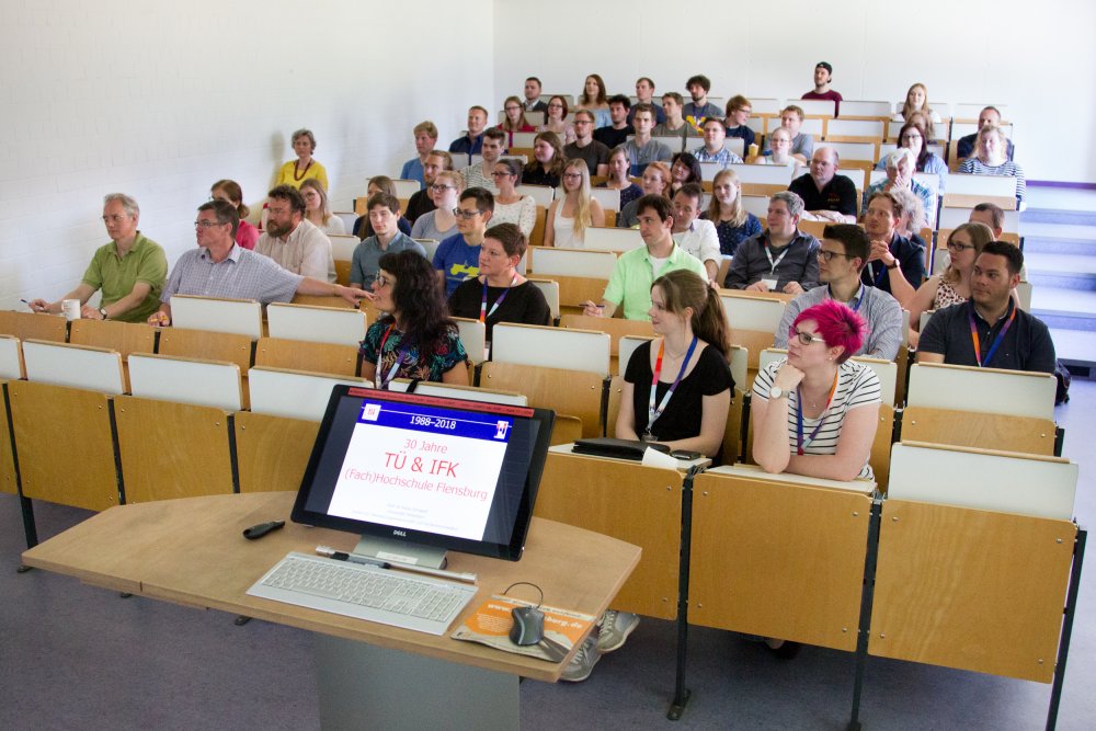 Lehrende und Studierende sitzen im Hörsaal, im Vodergrund ein Bildschirm, auf dem eine Präsentation zu erkennen ist.