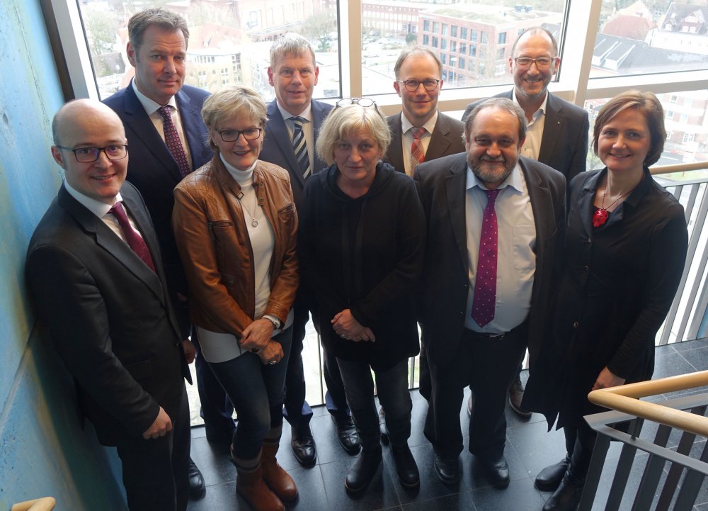 Eine Gruppe von Menschen, Männer in Anzügen und Frauen in Business-OUtfits, posieren in einem Treppenhaus vor einer Fensterwand.