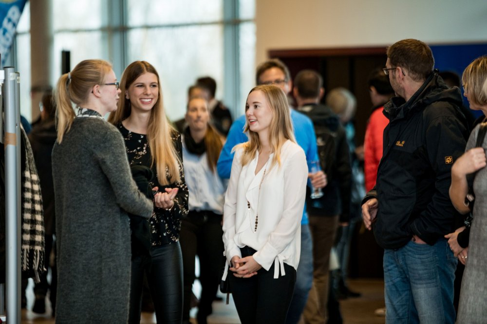 Menschen stehen im Gespräch im Foyer des audimax. In der Mitte des Bildes eine junge, lachende Frau.