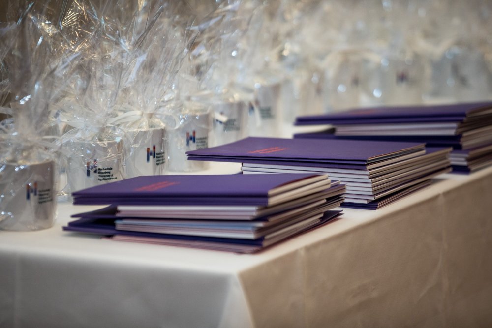 Urkundenmappen und in durchsichtiger Folie verpackte Hochschul-Tassen auf einem Weiß gedeckten Tisch.